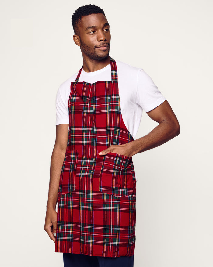 Two Petite Plume Adult Aprons in Imperial Tartan hang on a wooden rack with a patterned white dish towel. Perfect for holiday baking, a wooden spoon and spatula peek from an apron pocket against a warm, beige wall backdrop, creating an inviting scene for cozy cooking adventures.