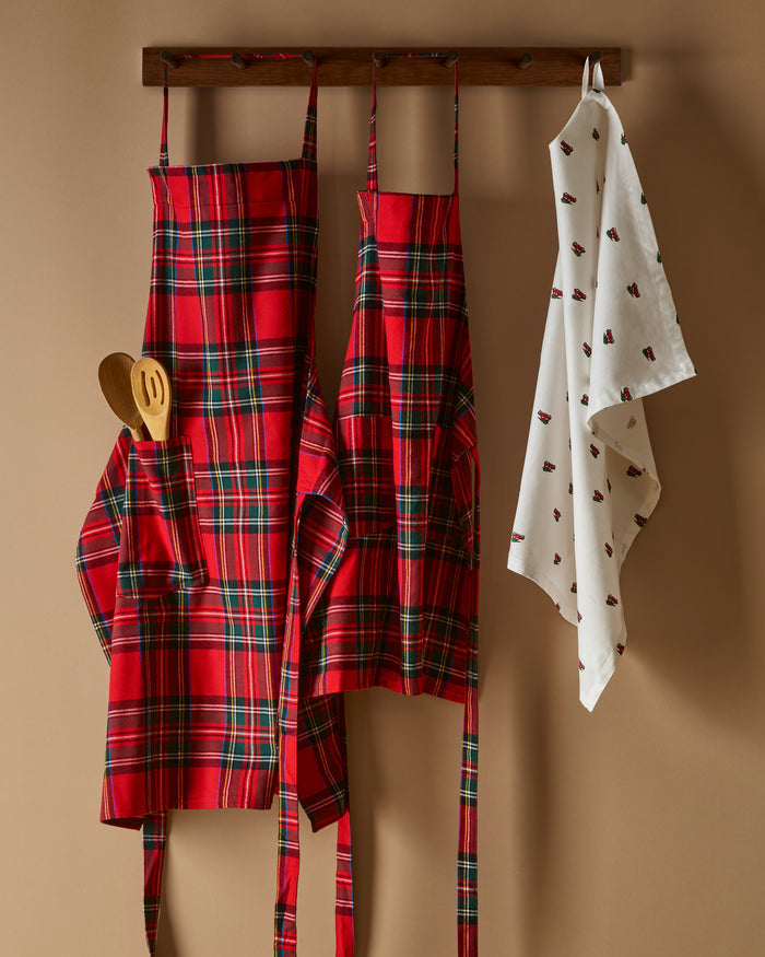 Two Petite Plume Adult Aprons in Imperial Tartan hang on a wooden rack with a patterned white dish towel. Perfect for holiday baking, a wooden spoon and spatula peek from an apron pocket against a warm, beige wall backdrop, creating an inviting scene for cozy cooking adventures.