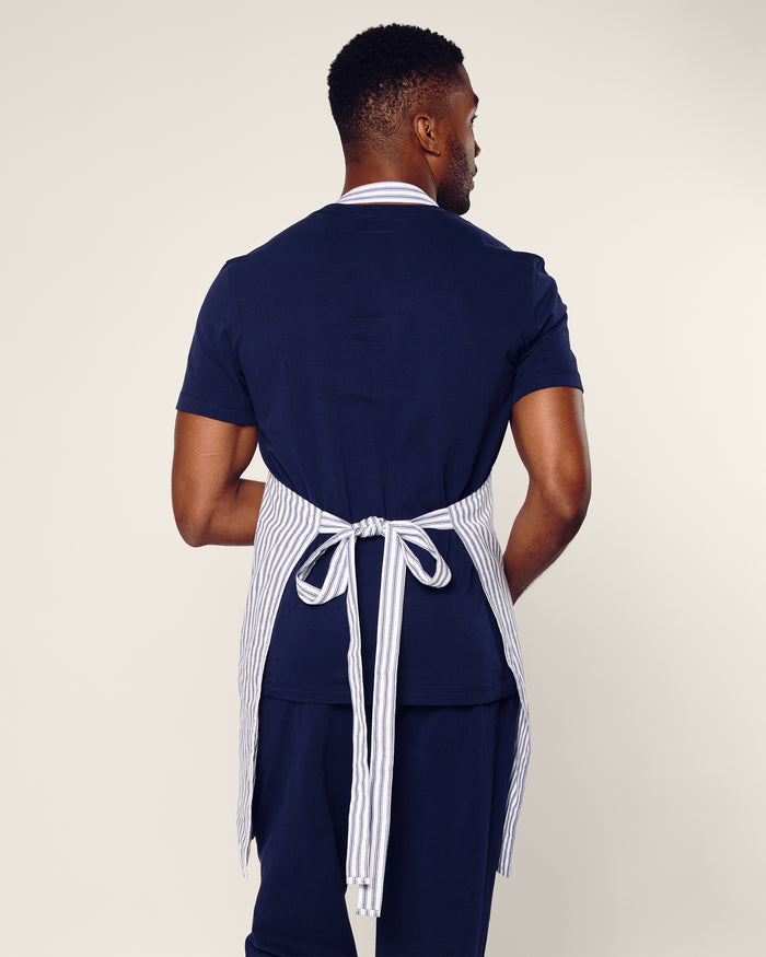 Against a neutral backdrop, a person in navy blue shirt and pants wears the Petite Plume Adult Apron in Navy French Ticking, exuding a holiday baking vibe.