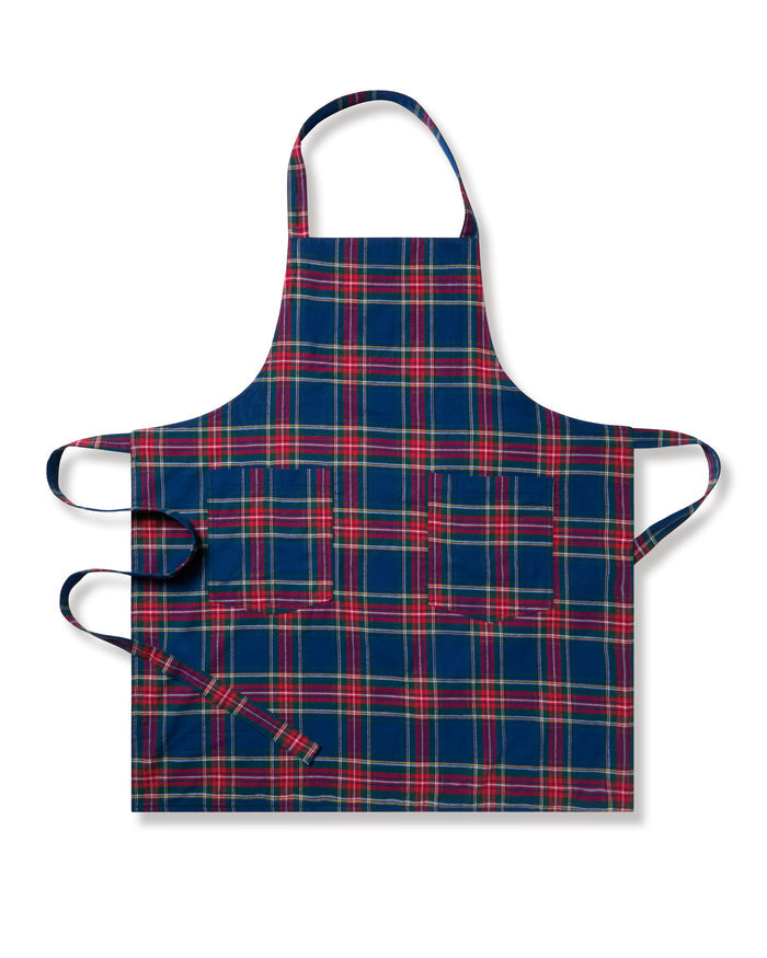 Two Adult Aprons in Windsor Tartan by Petite Plume, ideal for holiday baking, hang on a wooden rack beside a striped towel. One apron is labeled Dad, with a wooden spoon in its pocket. The background features a cozy warm beige wall.