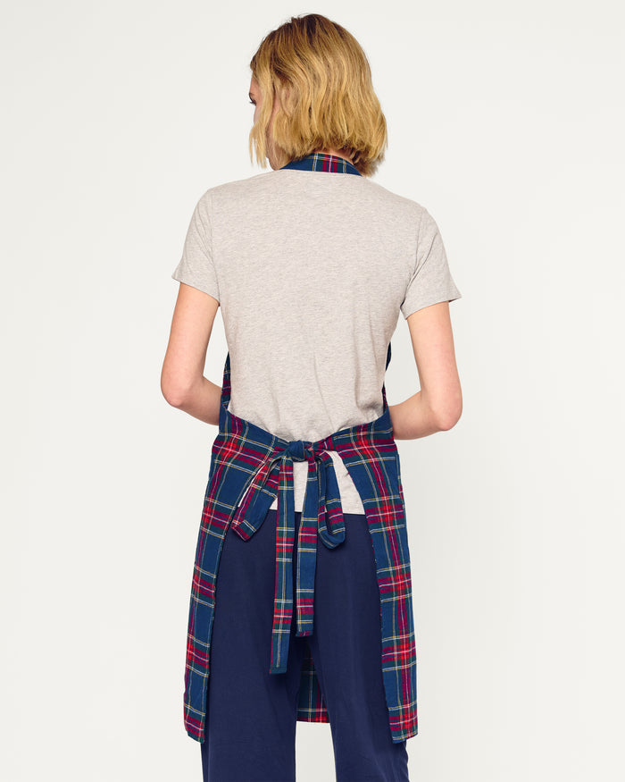Two Adult Aprons in Windsor Tartan by Petite Plume, ideal for holiday baking, hang on a wooden rack beside a striped towel. One apron is labeled Dad, with a wooden spoon in its pocket. The background features a cozy warm beige wall.
