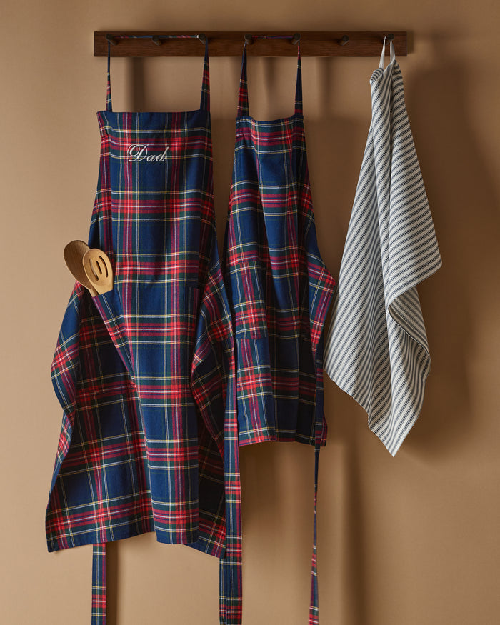 Two Adult Aprons in Windsor Tartan by Petite Plume, ideal for holiday baking, hang on a wooden rack beside a striped towel. One apron is labeled Dad, with a wooden spoon in its pocket. The background features a cozy warm beige wall.