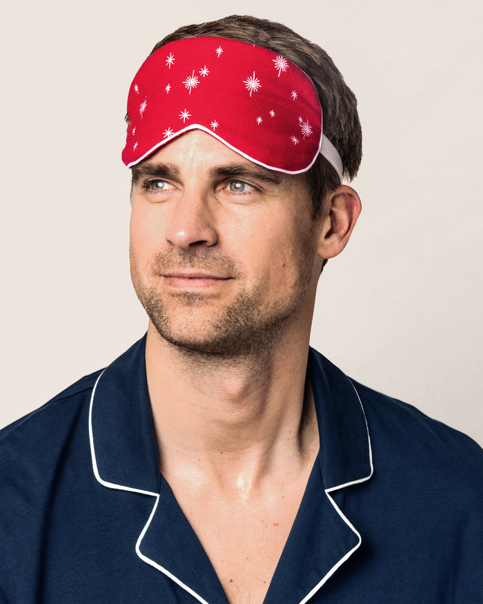 A man in navy pajamas with white trim and a red sleep mask featuring white stars looks aside against a simple backdrop, displaying Petite Plume’s Adults Flannel Sleep Mask in Starry Night, a perfect gift for any occasion.