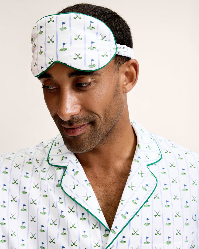 The Adult's Pima Sleep Mask in Fairway Stripe is a white sleep mask with green trim, decorated with golf clubs, balls, and putting greens. It features an adjustable strap and a small side tag.