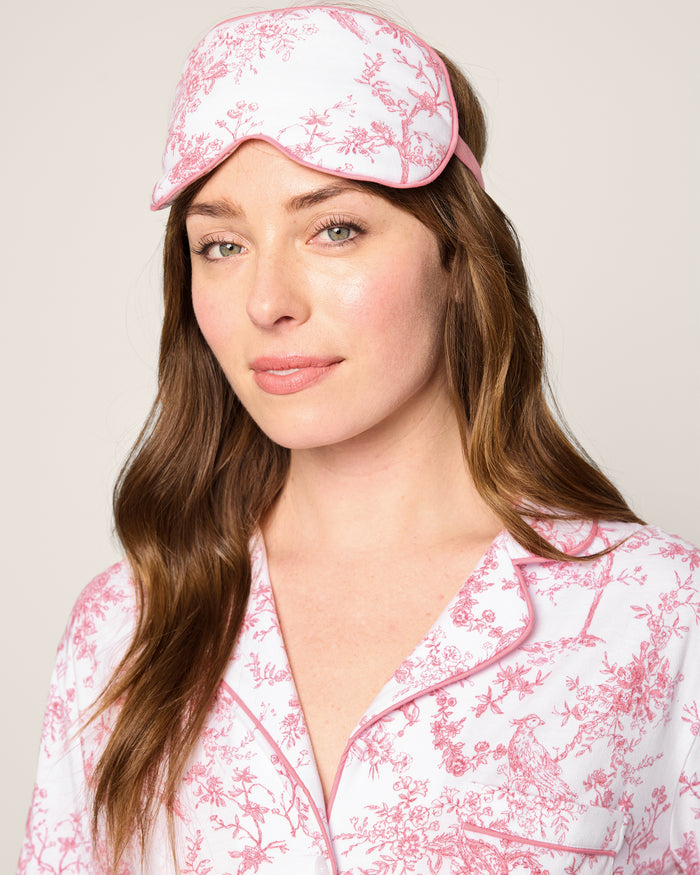 A woman with long brown hair wears a luxurious white and pink floral pajama set paired with the elegant Petite Plume Adults Pima Sleep Mask in Pink Timeless Toile adorned with botanical flowers on her forehead. She stands against a plain background, smiling softly.