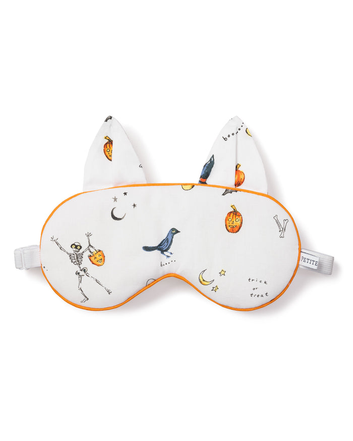 The Petite Plume Adults Twill Kitty Sleep Mask in Trick or Treat features a white design with orange trim, festive icons like a skeleton, bird, bats, pumpkins, stars, and trick or treat text. With cat ears and an adjustable strap, it adds spooky style to your sleep routine!.