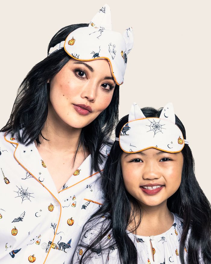 Two people wear matching Halloween pajamas with bat, pumpkin, and web prints alongside Petite Plumes Twill Kitty Sleep Masks in Trick or Treat. They smile against a plain background, ready for a cozy night in.