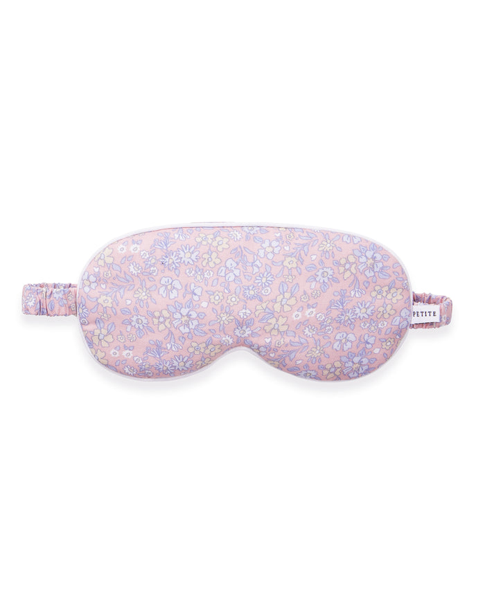 A woman with long brown hair wears the Adult's Twill Sleep Mask in Fleur Blush, lifting it slightly with one hand as she gazes off to the side with a relaxed expression.