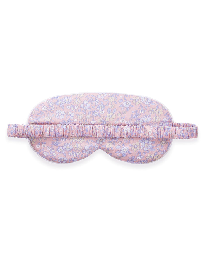 A woman with long brown hair wears the Adult's Twill Sleep Mask in Fleur Blush, lifting it slightly with one hand as she gazes off to the side with a relaxed expression.