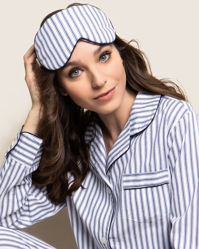 A person with long brown hair wears a pair of luxury dreamland blue and white striped pajamas. Resting atop their head is Petite Plumes Adults Twill Sleep Mask in Navy French Ticking. They are smiling slightly while sitting against a plain background.