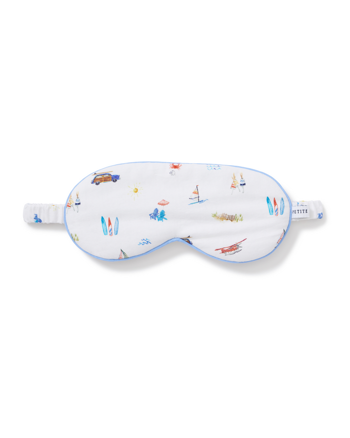 The Adult’s Twill Sleep Mask in Summer Nostalgia is a white mask with blue trim, featuring colorful beach-themed illustrations such as surfboards, boats, a jeep, and palm trees. It includes an elastic strap for a secure fit.