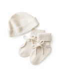 The Baby Cashmere Bootie & Hat Set in Ivory features cream-colored knitted booties with tied bows and a matching ribbed knit hat, both shown on a white background.