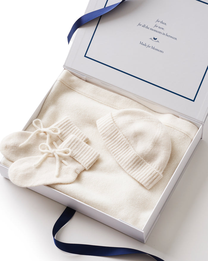 The Baby Cashmere Bundle in Ivory—featuring a folded knit blanket, matching baby hat, and bow-adorned booties—all made from soft cashmere, is arranged on a white background.