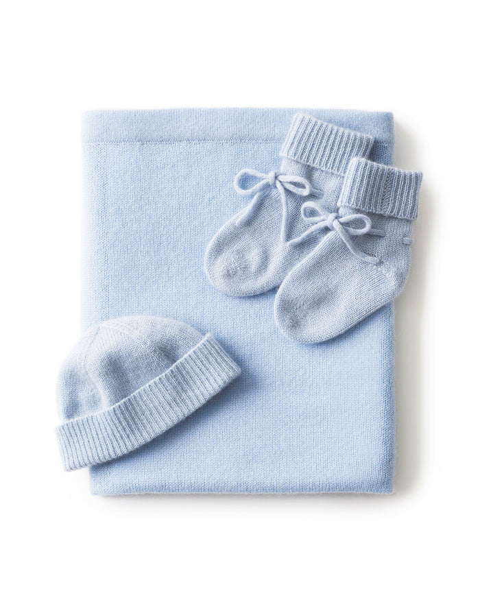 The Baby Cashmere Bundle in Light Blue features a neatly folded knit blanket, a matching baby hat, and knit booties with ribbons, all beautifully arranged on a white background.