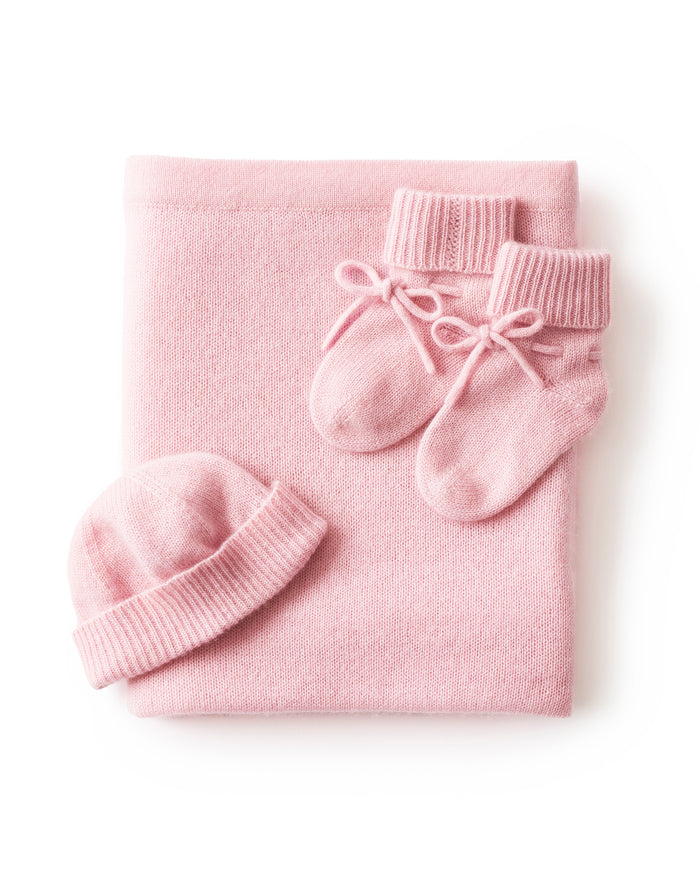 The Baby Cashmere Bundle in Rose features a neatly folded pink baby blanket, matching knit booties, and a ribbed knit hat, all displayed on a white background.