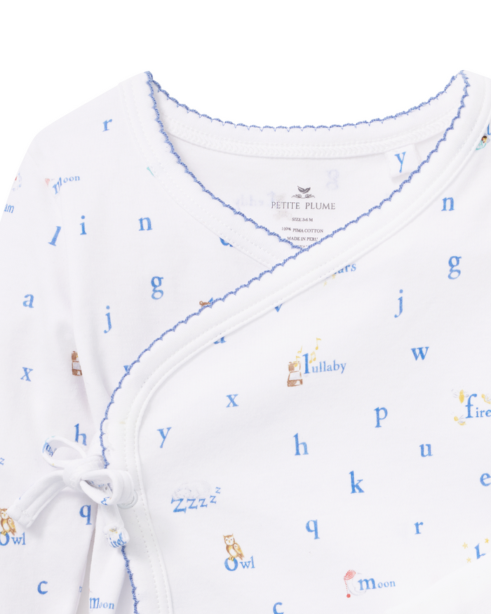 Close-up of the Baby Pima Wrap Set in Letters and Lullabies—a white baby onesie with blue trim, featuring alphabet letters, charming illustrations of animals and moons, and words like “lullaby” and “firefly.” The side tie closure is visible.