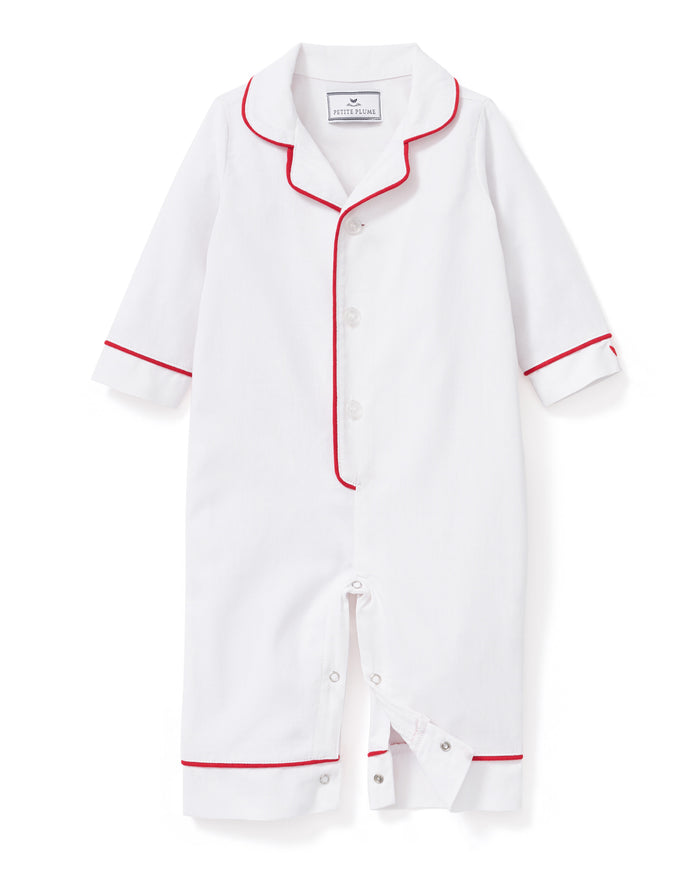 A toddler in Petite Plumes Babys Twill Romper in White with Red Piping stands against a plain background, looking up curiously with slightly tousled hair, as if just woken from sleepwear comfort.