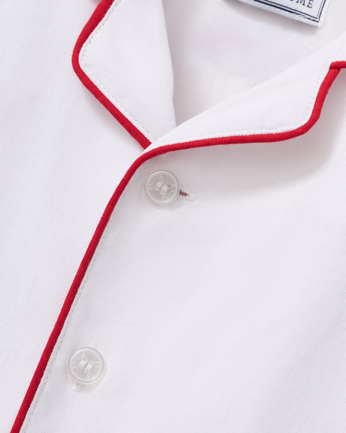 A toddler in Petite Plumes Babys Twill Romper in White with Red Piping stands against a plain background, looking up curiously with slightly tousled hair, as if just woken from sleepwear comfort.