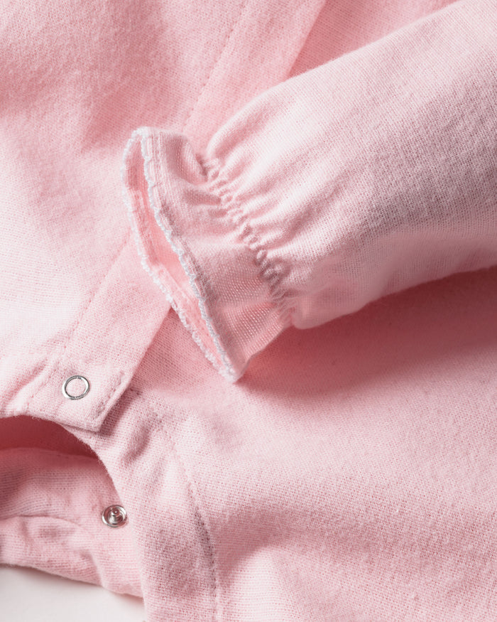 A close-up of Petite Plumes Babys Flannel Madeline Romper in pastel pink shows a brushed cotton flannel featuring long sleeves with elastic ruffle details and silver snap buttons on the front placket. The texture is soft and fleece-like, perfect for childrens sleepwear.