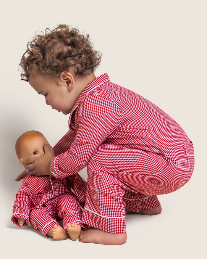 A toddler with curly hair wears Petite Plumes Babys Flannel Romper in Red Mini Gingham and plays with a matching doll, gently touching its face. They are set against a light, plain background that enhances the charming scene.