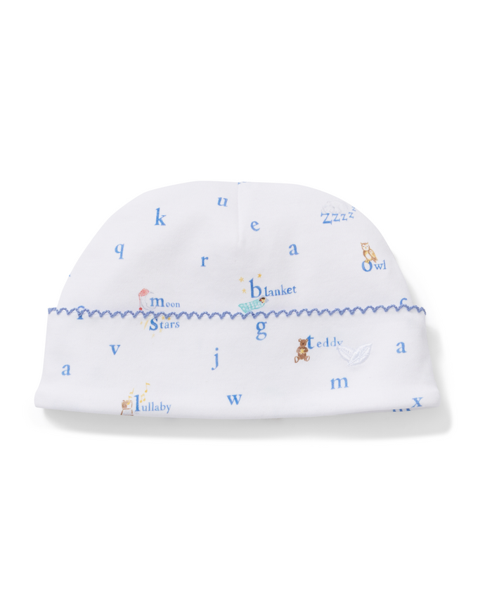 Introducing the Baby’s Pima Hat in Letters and Lullabies—a white baby hat featuring a blue zigzag trim, playful animal illustrations, stars, teddy bears, scattered blue letters, whimsical motifs, and sweet words like “blanket.”.
