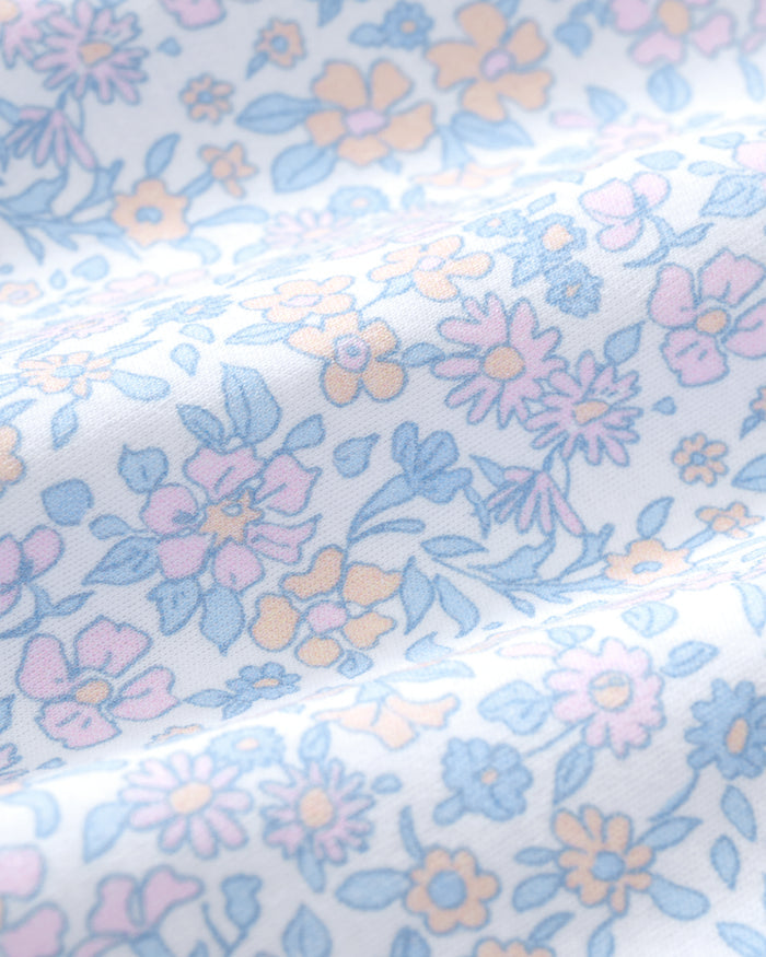 The Baby's Pima Isla Play Romper in Fleur Blanche features light blue and white florals, short ruffled sleeves, and elastic leg openings, shown laid flat on a white background.
