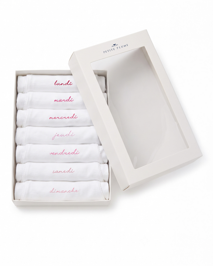 A set of seven white Baby’s Pima Long Sleeve Onesies from the “French Lessons Pink” collection, each embroidered in pink with a day of the week in French, neatly arranged in an open cream box with a clear lid.