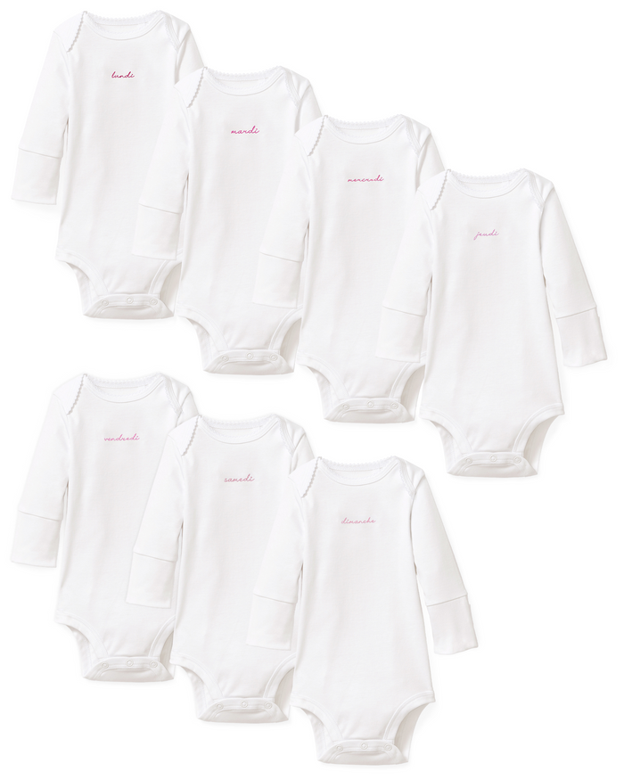 Seven white long-sleeve Baby’s Pima Onesies from the French Lessons Pink set are shown, each featuring a day of the week embroidered in pink cursive Spanish lettering on the chest.
