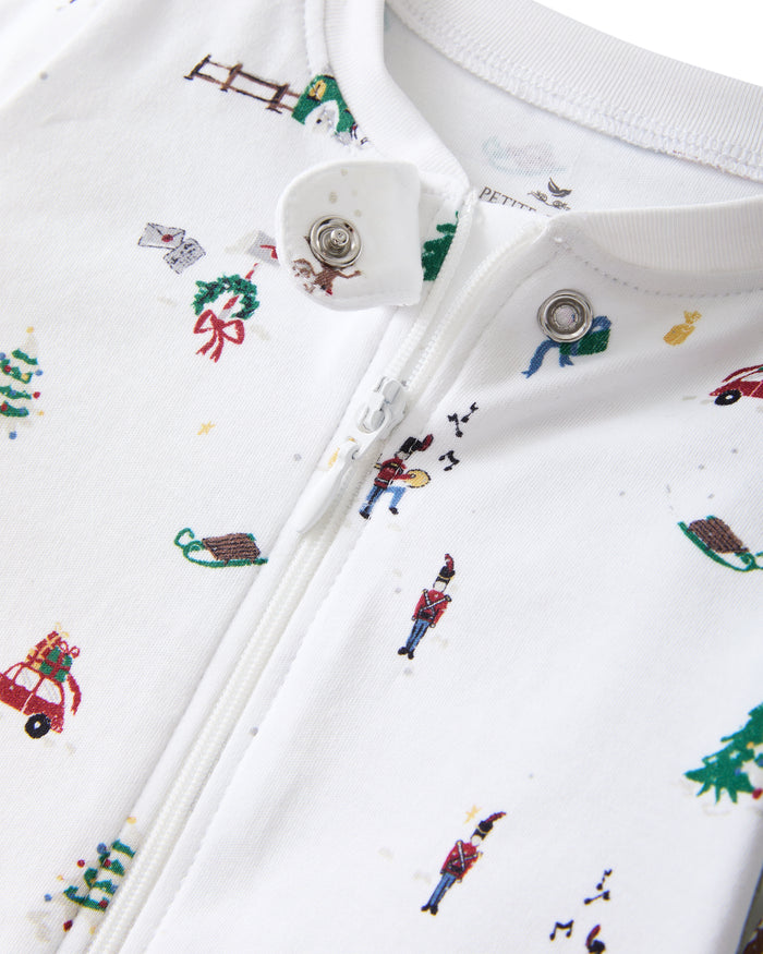 The Baby's Pima Romper in Holiday Town is a white footed sleeper with a diagonal zipper, featuring colorful prints of Christmas trees, reindeer, cars, snowmen, fences, and hearts.