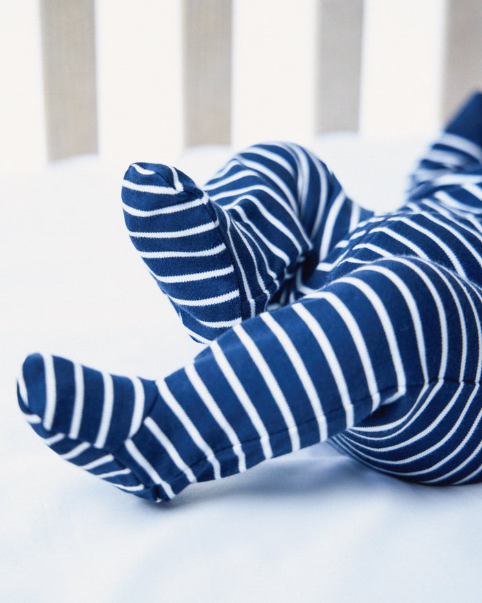 A baby rests in a crib, clad in Petite Plumes Babys Pima Romper in Navy Stripe. The legs and part of the body peek through the slats, highlighting its cozy Pima cotton comfort.