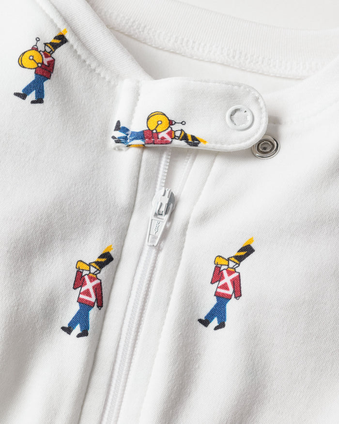 The close-up displays Petite Plumes Babys Pima Romper in Toy Soldier, crafted from soft Pima cotton. It features a zipper and snap button with a charming print of toy soldiers in red jackets, black hats, and carrying instruments repeatedly adorning the fabric.