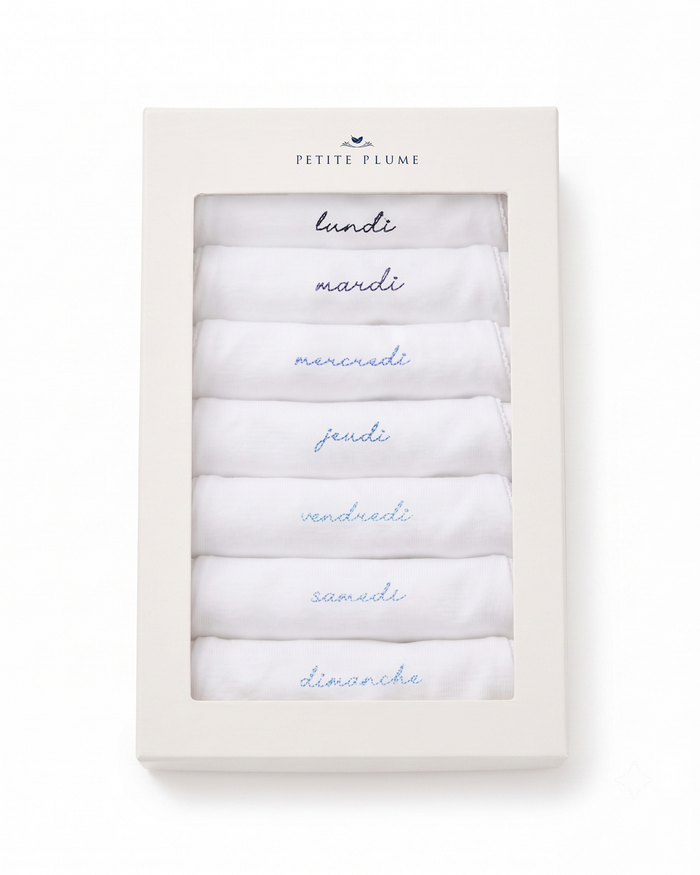 A boxed set labeled Petite Plume, containing seven white cloth Baby’s Pima Short Sleeve Onesies in French Lessons Blue, each embroidered with a day of the week in French—lundi, mardi, mercredi, jeudi, vendredi, samedi, dimanche—arranged in order.