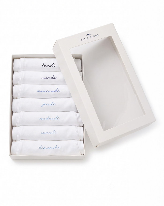A white box with a clear lid showcases the Baby’s Pima Short Sleeve Onesie Set in French Lessons Blue, featuring seven neatly folded white onesies, each labeled with a day of the week in French: lundi, mardi, mercredi, jeudi, vendredi, samedi, and dimanche.