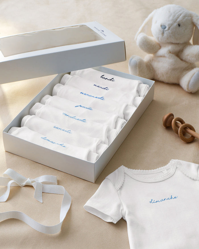A box featuring the Baby’s Pima Short Sleeve Onesie Set in French Lessons Blue—five white baby onesies marked with days of the week in French—alongside a plush bunny, wooden rattle, decorative ribbon, and a special Sunday (“dimanche”) onesie with blue embroidery, all displayed on a beige surface.