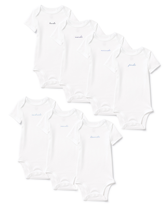 Seven white Baby’s Pima Short Sleeve Onesies from the French Lessons Blue set are arranged in two rows, each featuring a day of the week written in cursive on the chest in light blue or gray text.