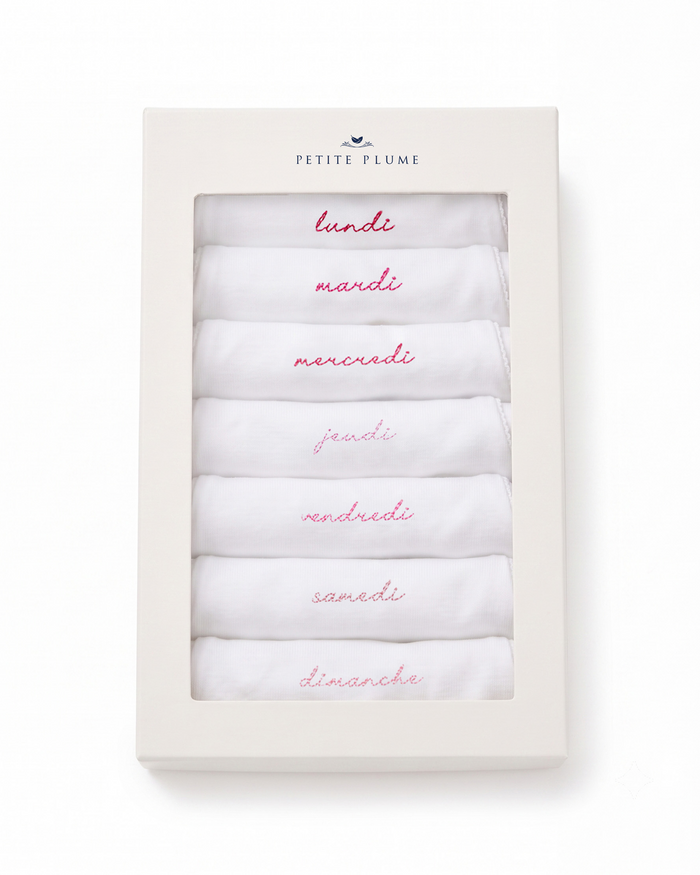 A box labeled Petite Plume at the top contains seven folded white Baby’s Pima Short Sleeve Onesies from the French Lessons Pink set, each embroidered in red with a day of the week in French—lundi, mardi, mercredi, jeudi, vendredi, samedi, and dimanche.