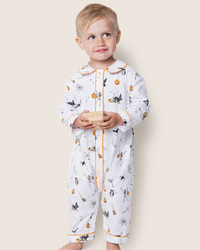 A child wearing Petite Plumes Babys Twill Cambridge Romper in Trick or Treat, decorated with pumpkins, witches, and cats, holds a small wooden block. The flame-retardant onesie with orange trim is set against a plain light background as the child gazes upward with a slight smile.
