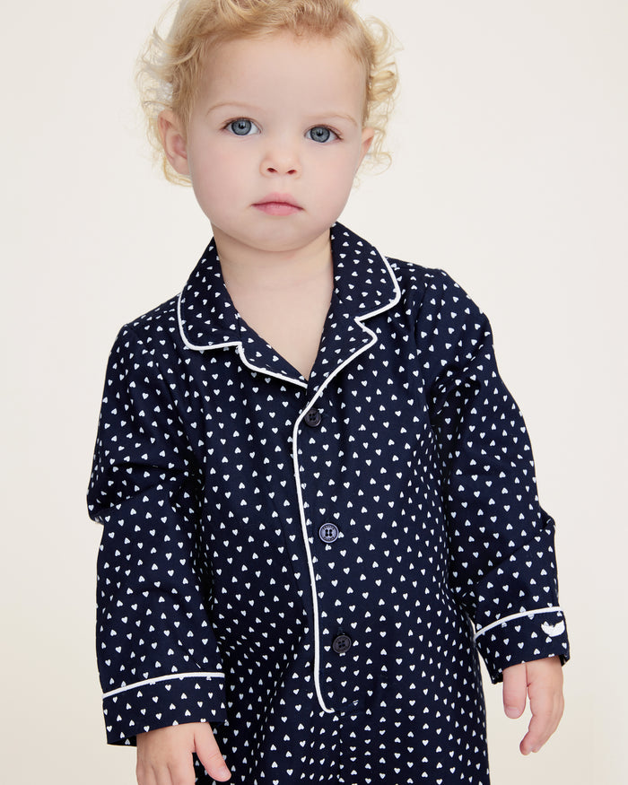 The Baby's Twill Classic Romper in Midnight Hearts is a navy blue onesie with long sleeves and legs, featuring a white heart pattern and white piping on the collar, cuffs, and front.