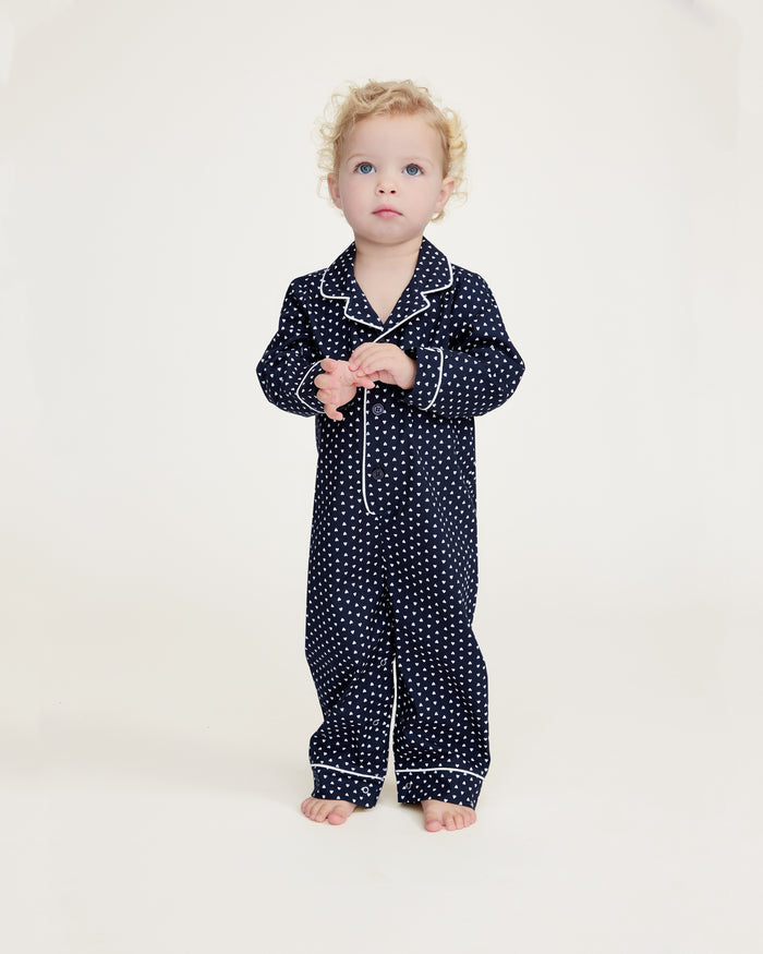 The Baby's Twill Classic Romper in Midnight Hearts is a navy blue onesie with long sleeves and legs, featuring a white heart pattern and white piping on the collar, cuffs, and front.