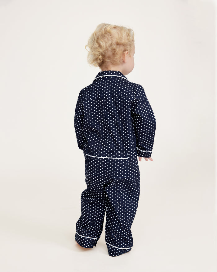 The Baby's Twill Classic Romper in Midnight Hearts is a navy blue onesie with long sleeves and legs, featuring a white heart pattern and white piping on the collar, cuffs, and front.