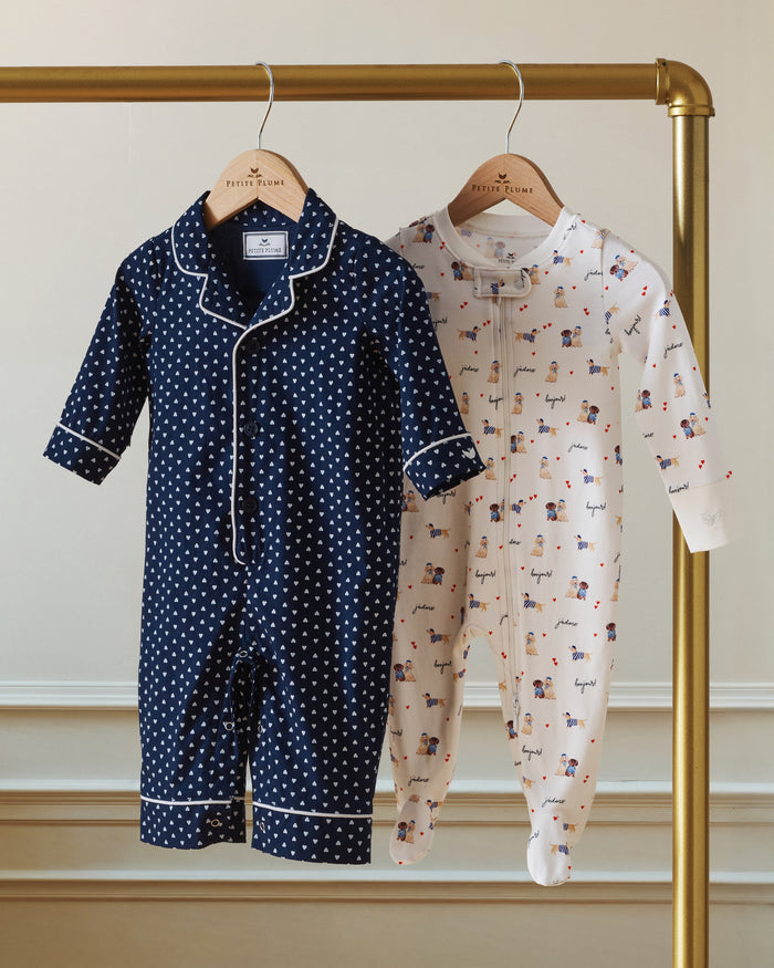 The Baby's Twill Classic Romper in Midnight Hearts is a navy blue onesie with long sleeves and legs, featuring a white heart pattern and white piping on the collar, cuffs, and front.