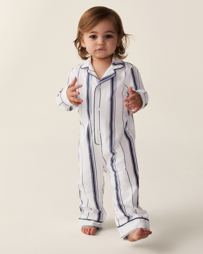 A toddler stands barefoot on a plain light background, wearing the Baby’s Twill Classic Romper in Parlor Stripe—white with dark vertical stripes—and gazes forward with a curious expression.