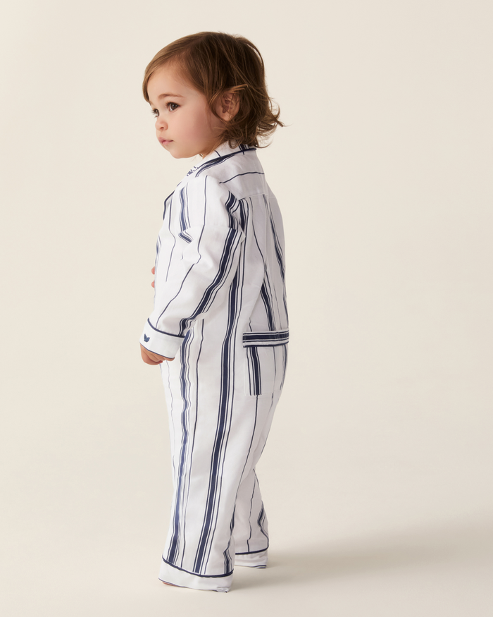 A young child with brown hair stands sideways against a plain light background, wearing the Baby’s Twill Classic Romper in Parlor Stripe—a white romper featuring vertical navy blue stripes.