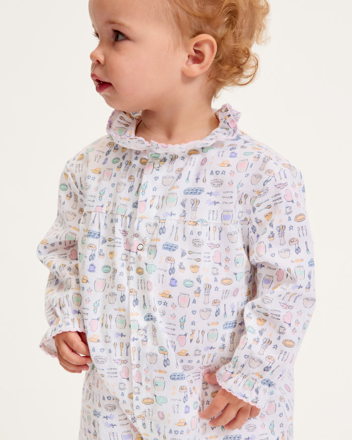 The Baby's Twill Madeline Romper in Baking Spirits Bright features long sleeves, a ruffled collar and cuffs, pastel kitchen print on white, front buttons, and snap closures at the legs for easy dressing.