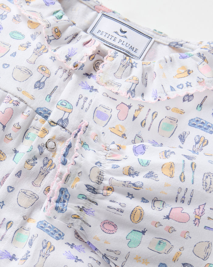 The Baby's Twill Madeline Romper in Baking Spirits Bright features long sleeves, a ruffled collar and cuffs, pastel kitchen print on white, front buttons, and snap closures at the legs for easy dressing.