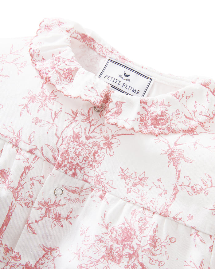 The Baby's Twill Madeline Romper in Pink Timeless Toile features long sleeves, pink toile floral and tree patterns, ruffled cuffs and collar, and convenient inseam snaps for easy diaper changes.