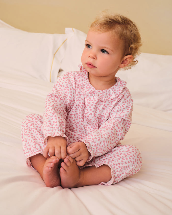 The Baby's Twill Madeline Romper in Sweethearts is a pink and white long-sleeve romper featuring a ruffled collar and cuffs, adorned with a small pink heart pattern.