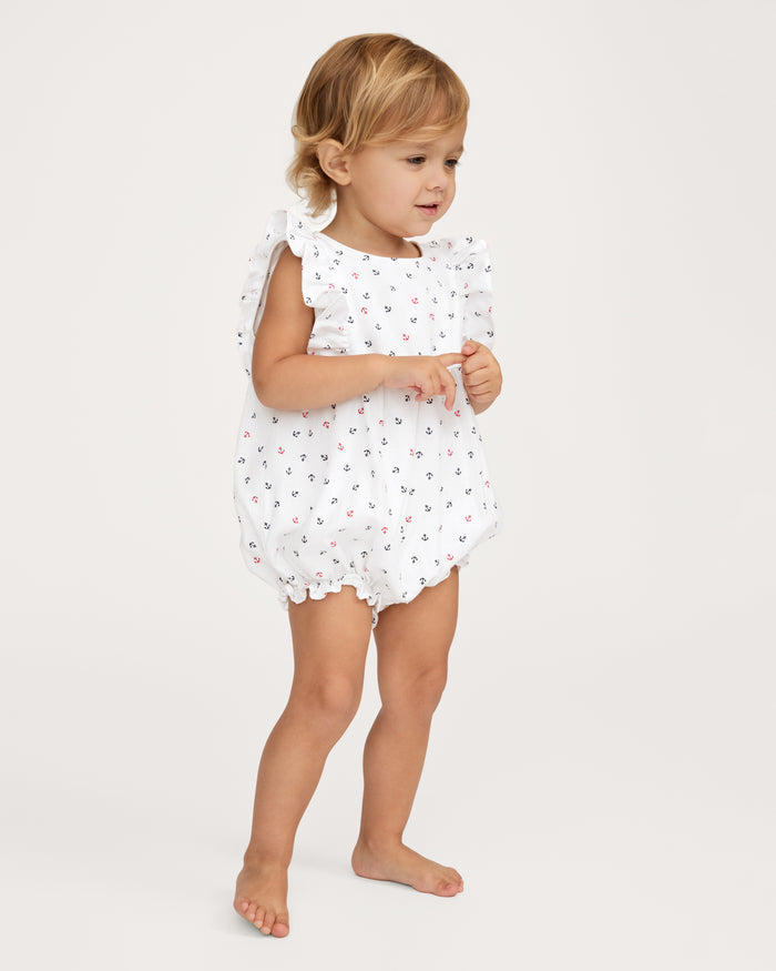 A young child with light brown hair is wearing the Baby’s Twill Ruffled Romper in Anchors Away White, a sleeveless white romper adorned with small anchors and hearts. The child stands barefoot on a plain, light background, gazing slightly to the left.