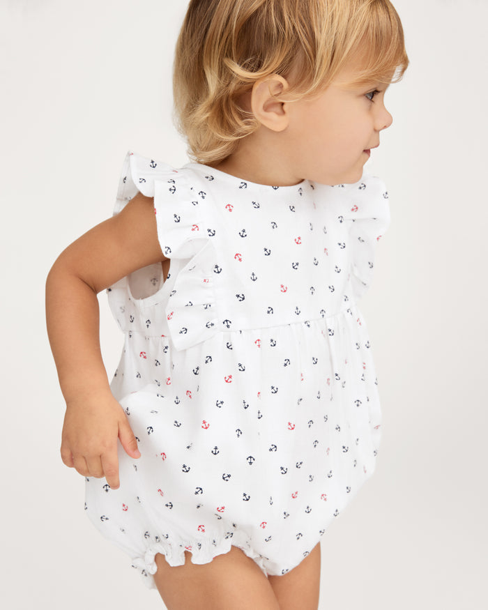 A toddler wearing the Baby’s Twill Ruffled Romper in Anchors Away White, featuring small anchor prints and ruffled sleeves, looks to the right against a plain background.