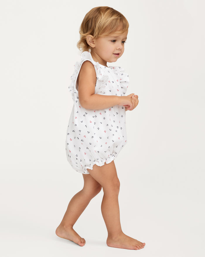 A young child with light brown hair wears the Baby’s Twill Ruffled Romper in Anchors Away White, a sleeveless white piece adorned with small anchor prints. The child is barefoot and walking, hands clasped, against a plain white background.